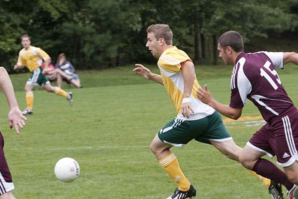 Two soccer players are chasing the ball during a game on a grass field. One player in a yellow and green jersey is ahead, while another player in a maroon jersey is close behind. Two more players and a couple of spectators are visible in the background.