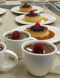 Cups of chocolate mousse topped with raspberries and plates of caramel flan garnished with blueberries and raspberries are arranged on a light-colored surface.