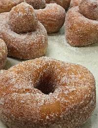 Close-up view of several sugar-coated, doughnut-shaped pastries and a few doughnut holes on a white surface. The pastries are golden brown with a light dusting of sugar, showcasing a soft texture.