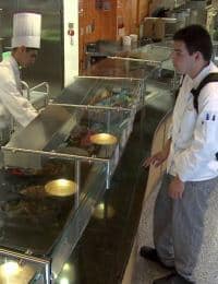 A chef, wearing a tall white hat, and a customer stand at a seafood counter. The chef is behind the counter, possibly preparing or selecting seafood. The customer, wearing a backpack, is on the other side, observing or making a decision.