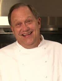 A man in a white chef's coat is smiling warmly at the camera. He has short hair and is standing in a kitchen setting.