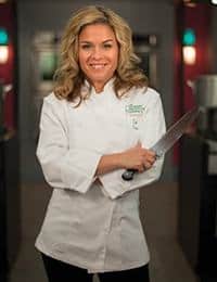 A smiling person with wavy, blonde hair stands confidently in a kitchen, wearing a white chef's coat and holding a large kitchen knife. The background is softly lit with colorful, blurred lights and kitchen equipment visible in the distance.