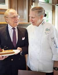 An elderly man in a suit and a younger person in a chef's uniform smile at each other. The elderly man holds a plate with a slice of cake while standing in a modern kitchen with wooden cabinets and large windows in the background.