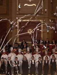 A group of chefs in white uniforms and hats, alongside formally dressed staff, stand in a grand room with a high ceiling and ornate decorations. They are celebrating, with glittering streamers and confetti falling from above. In the background, a few people observe the scene.