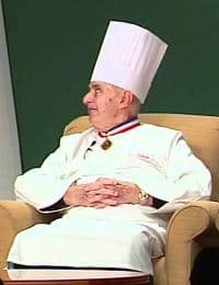 An elderly chef wearing a tall white chef's hat and traditional white chef's coat adorned with medals and decorations is seated on a beige chair, looking to his right. His hands are clasped in his lap, and he exudes an air of distinguished professionalism.