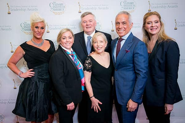 A group of six people, three men, and three women, dressed in formal attire, stand together smiling for a picture at the Culinary Institute of America Leadership Awards event. The backdrop features the event's name and logos.