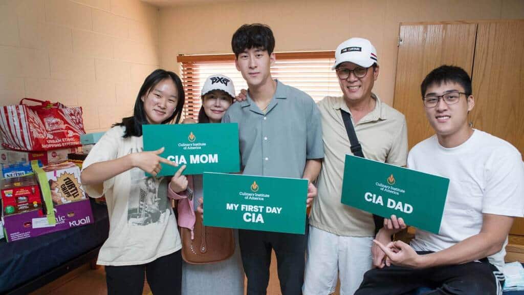 A student poses with family members in a room, all holding signs. The student’s sign reads "My First Day CIA," while others read "CIA Mom" and "CIA Dad." They are smiling and standing next to stacked boxes and supplies, suggesting the student’s move-in day.