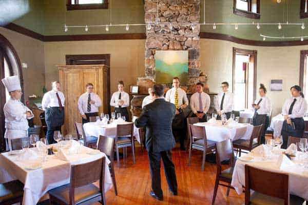 A group of restaurant staff stand attentively in a formal dining room as a manager or instructor, dressed in a suit, addresses them. The room features wooden floors, neatly set tables, an exposed stone wall, and soft overhead lighting.