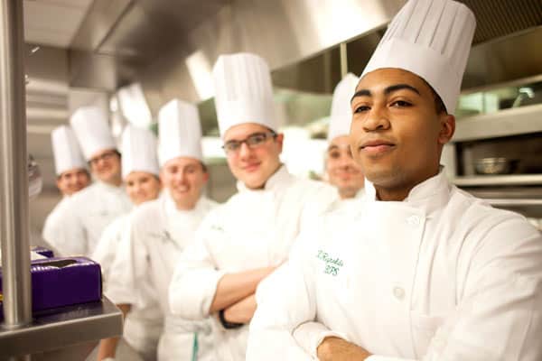 A group of chefs in white uniforms and tall white hats stand in a commercial kitchen. The chef in the foreground has arms crossed and looks forward confidently, while the others in the background smile and also stand with crossed arms.