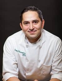 A chef with short dark hair and a slight smile is wearing a white chef's jacket. The text on the jacket reads "Cory D. Chartrand-Zwaschka" and has the initials "CFS." The background is black, providing a clear contrast to his attire.