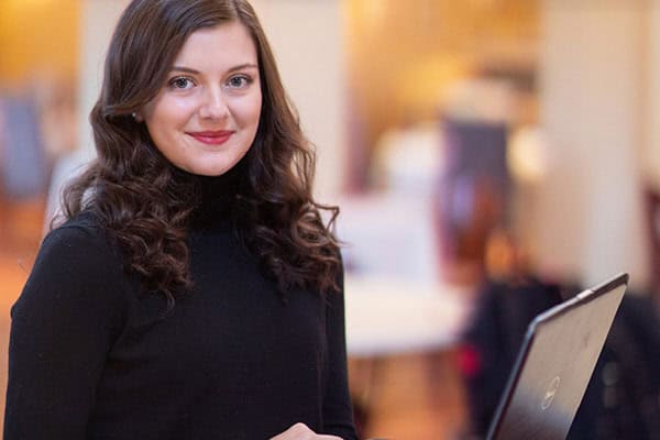 A woman wearing a black long-sleeved top is standing and smiling at the camera. She has long, wavy brunette hair and is using a laptop. The background is softly blurred, with warm tones and various lights.