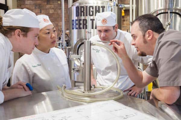 Four people in a brewery laboratory are wearing white coats and hats, closely observing a brewing apparatus. One person in a gray shirt explains something while the others look on intently. Large metal brewing tanks labeled "BRIGHT" are in the background.