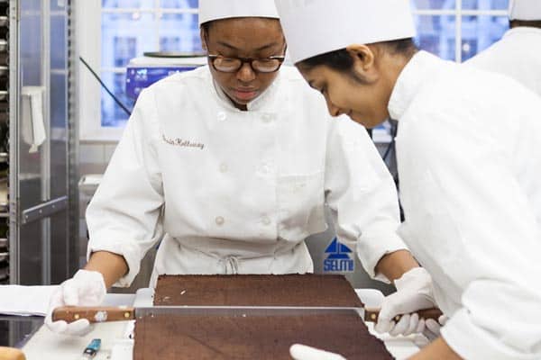 Two chefs in white uniforms and hats are working in a kitchen, focusing on a large rectangular chocolate slab. One chef uses a ruler for precise measurements, while the other observes closely. Cabinets and kitchen equipment are visible in the background.