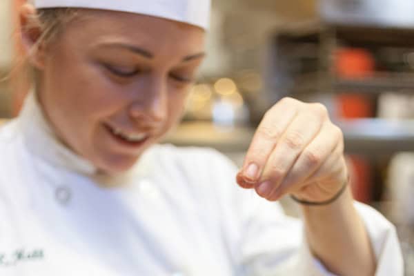 A chef in a white uniform and chef's hat is focusing while sprinkling a pinch of seasoning into a dish. The background is blurred, showing kitchen equipment. The chef is smiling gently, conveying her passion and dedication to her culinary craft.