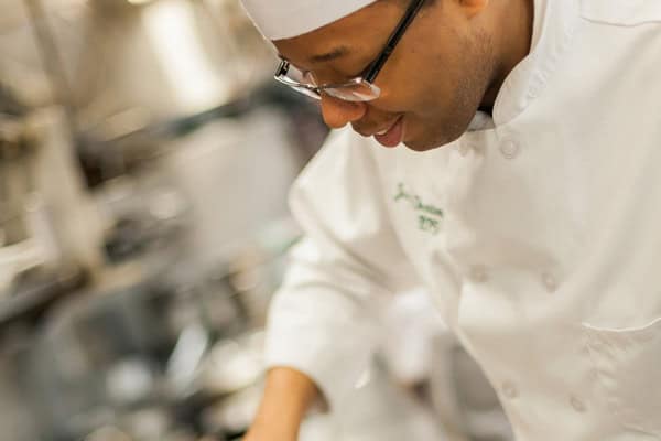 A chef wearing glasses and a white chef's uniform with a hat is focused on preparing food in a kitchen. The background is blurred, emphasizing the chef's concentration and the professional kitchen setting.