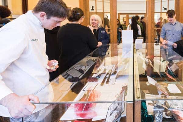 A man dressed in a chef's uniform looks at items displayed in a glass case. Several people are gathered around another display in the background, engaged in conversation. The setting appears to be an exhibit or a museum.