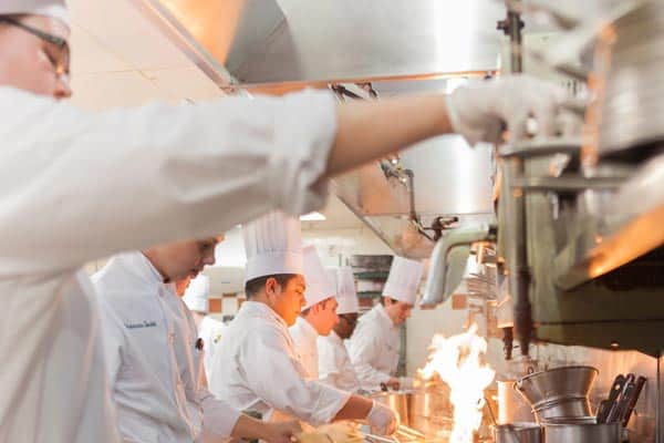 Chefs in white uniforms and tall hats are focused on cooking in a busy, professional kitchen. Flames can be seen rising from a stovetop as they work with various utensils and ingredients.