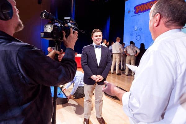 A man in a bow tie stands on a stage, smiling as he is interviewed by another person holding a microphone. A cameraman films the interview. In the background, other people are present, and a projector screen displays a graphic.