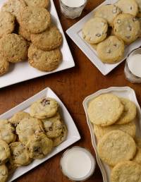 A wooden table with four plates of cookies, including chocolate chip and other varieties. Three glasses of milk are placed around the plates, enhancing the cozy, inviting atmosphere.