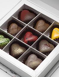 A white box containing nine heart-shaped chocolates of various colors and patterns. There are brown, red, yellow, and green chocolates with intricate surface designs. Each chocolate is placed in an individual compartment within the box.