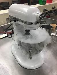 A white stand mixer on a countertop is surrounded by a cloud of vapor emanating from its mixing bowl. The mixer is plugged in, with some kitchen utensils and containers in the background.