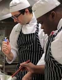 Two chefs in white uniforms and striped aprons work together in a commercial kitchen. One chef is seen holding a whisk, while the other focuses on stirring a pot. Both wear white hats and appear to be engaged in food preparation.