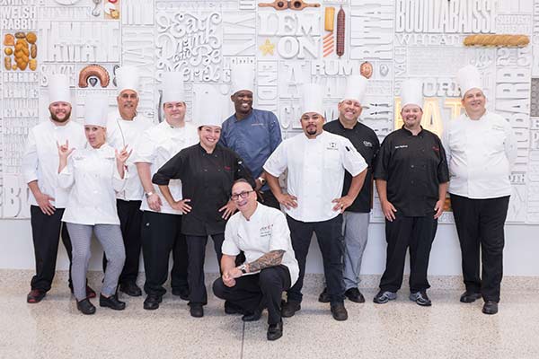 A group of ten chefs, both men and women, are standing and posing in front of a decorative wall with culinary-themed words and images. They are all wearing their chef uniforms, consisting of white and black jackets. Some are smiling and others gesture playfully.