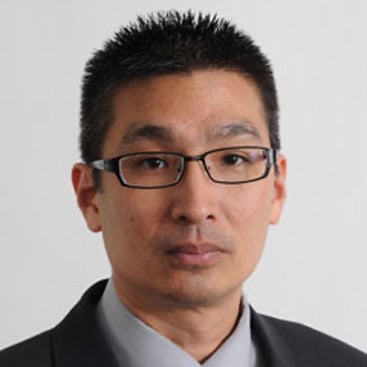 A person with short, spiked black hair and rectangular glasses is wearing a dark suit, light dress shirt, and tie. They are posed against a plain white background, looking directly into the camera with a neutral expression.