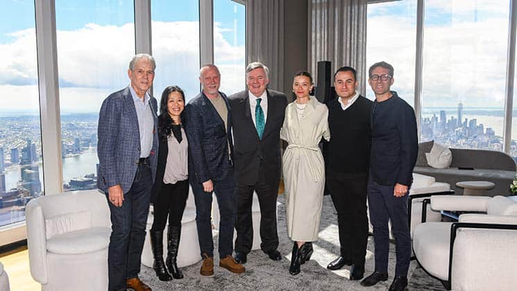 Seven people are standing together in a modern, high-rise room with large windows, offering a scenic view of a city skyline and river. The group is a mix of men and women dressed in business casual attire, smiling for the photo.