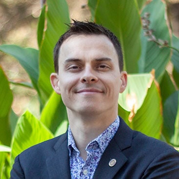 A person with short dark hair is smiling with arms crossed, wearing a dark blazer over a patterned shirt. They are standing in front of large green leaves, possibly outdoors.