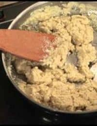 A close-up of a pan on a stovetop containing a thick, lumpy mixture being stirred with a wooden spatula. The mixture appears to be in the process of cooking, with an uneven texture and a light brown color.