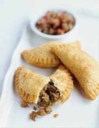 Three golden-brown empanadas are on a white plate, one of them broken open to reveal a savory meat filling. In the background, there is a small dish of what appears to be salsa.