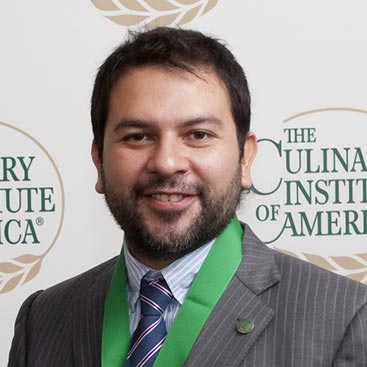 A man with short dark hair and a beard smiles at the camera. He is wearing a suit, a striped tie, and a green ribbon around his neck. The backdrop features the logo and text "The Culinary Institute of America.