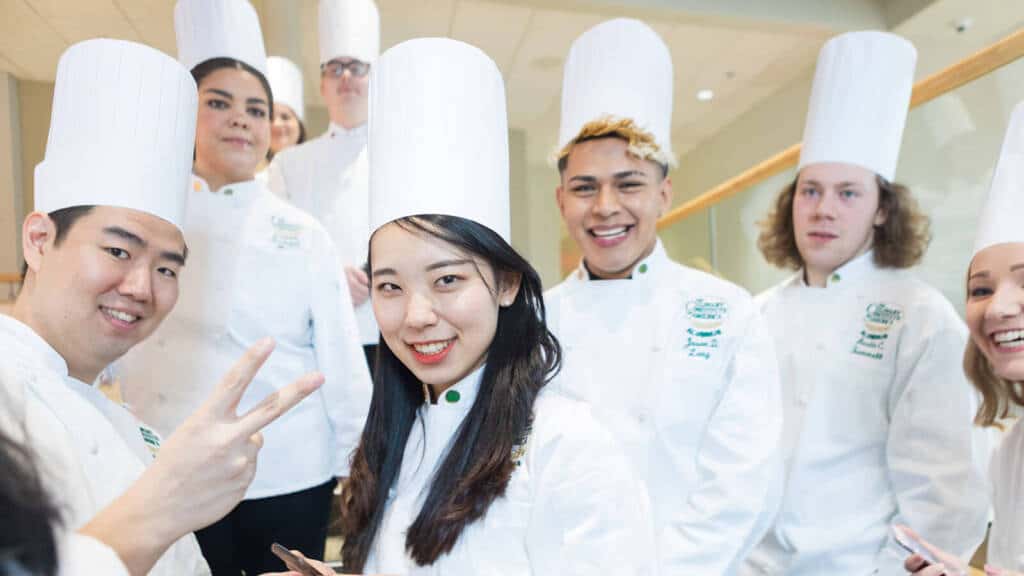 A group of culinary students in white uniforms and tall chef hats stand together, smiling at the camera. One student in the front is making a peace sign with his fingers. They appear to be in a bright, indoor setting, perhaps a culinary school or kitchen.