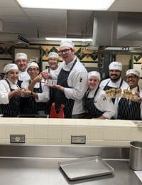 A group of chefs and culinary students in white uniforms and chef hats stand smiling in a commercial kitchen. They are holding dishes and utensils, appearing happy and proud of their work, with various kitchen equipment and a large stainless steel counter in the foreground.