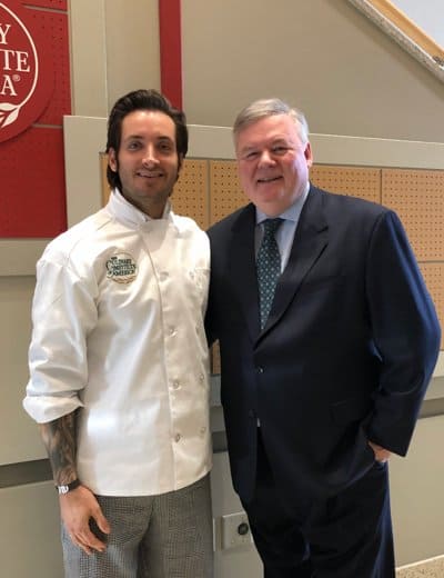 Two men stand side by side, smiling. The man on the left wears a white chef's jacket and checkered pants, while the man on the right wears a navy blue suit with a white shirt and dotted tie. They are in an indoor setting with a red emblem on the wall behind them.