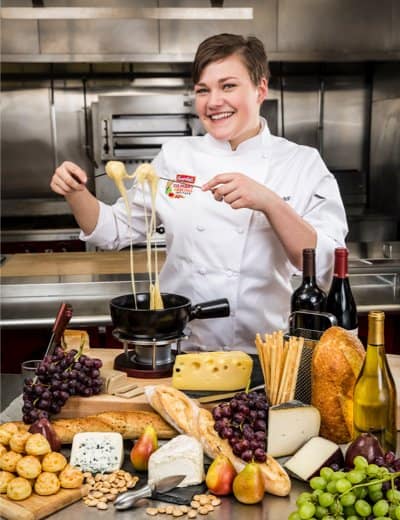 A smiling chef in a white uniform is holding a fork with melted cheese over a fondue pot. The table in front is filled with an assortment of cheeses, bread, grapes, pears, nuts, and bottles of wine. The background features a professional kitchen.