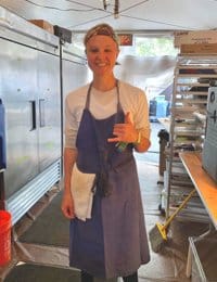 A smiling person dressed in a white shirt and blue apron stands in a kitchen, gesturing with a "shaka" hand sign. The spacious kitchen has a large refrigerator, shelves, and various equipment in the background.