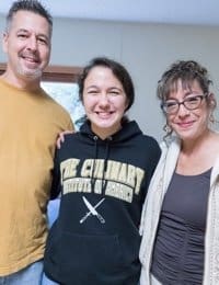 A smiling group stands together indoors. From left to right: a man in a yellow shirt, a young woman in a black hoodie with "The Culinary Institute of America" written on it, and a woman with glasses wearing a beige sweater over a brown top.