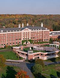 Aerial view of a large, red-brick, multi-story building with white accents, surrounded by landscaped gardens and pathways. The building features columns, arches, and a clock tower. It is set against a backdrop of trees with autumn foliage.