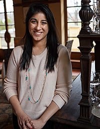 A woman with long dark hair, wearing a light sweater and a beaded necklace, stands indoors smiling at the camera. She is positioned in front of a wooden structure, with soft natural light coming through the windows in the background.