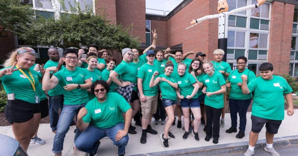 A diverse group of people, all wearing green T-shirts, stand together in front of a brick building, enthusiastically flexing their biceps and smiling. A large puppet figure with outstretched arms is visible in the background near the building.