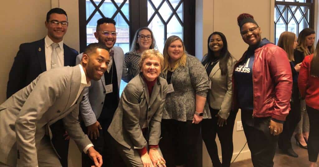 A group of eight people, dressed in business attire, are smiling and posing for a photo in a room with windows. They stand close together, some slightly leaning forward. The setting appears to be professional, possibly a conference or networking event.