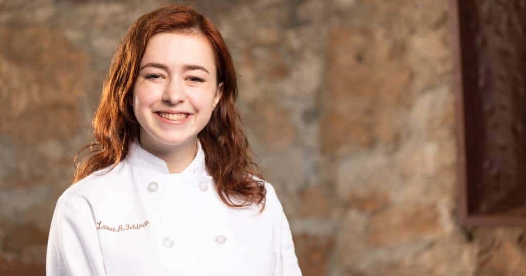 A young woman with long, wavy hair and a bright smile stands against a stone wall background. She is wearing a white chef's coat.