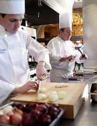 Two chefs in white uniforms and hats working in a bright kitchen. The male chef in the foreground is chopping onions at a wooden cutting board. The chef in the background supervises, holding a utensil with blurred ingredients in the foreground.