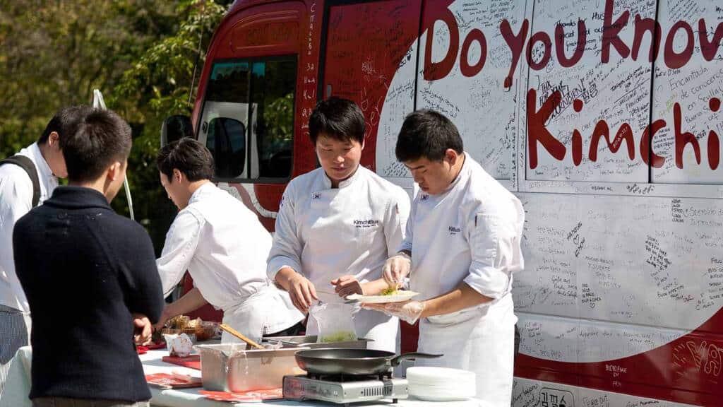 A group of chefs in white uniforms prepare food at an outdoor event in front of a food truck covered with handwritten notes and the words "Do you know Kimchi?" A few people stand nearby watching the chefs work. Cooking equipment and ingredients are on the table.