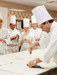 A group of culinary students in white uniforms and tall white chef hats observe a chef demonstrating a technique at a countertop in a kitchen. The chef at the foreground is handling a piece of food with a tool, while students take notes.