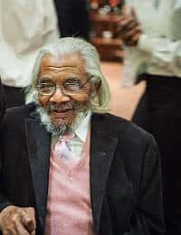 An elderly man with long white hair and a beard, wearing glasses, a white shirt, a pink pullover, and a black jacket, smiles while seated. Other people in formal attire stand around him in the background.