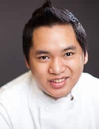 A smiling person with short, dark hair is wearing a white chef's coat. The background is blurred and dark, making the subject stand out clearly.