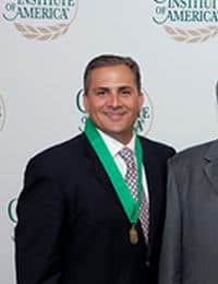 A man wearing a dark suit, white shirt, and patterned tie with a green ribbon and medal around his neck stands in front of a backdrop with the text "Institute of America." He is smiling and facing the camera.